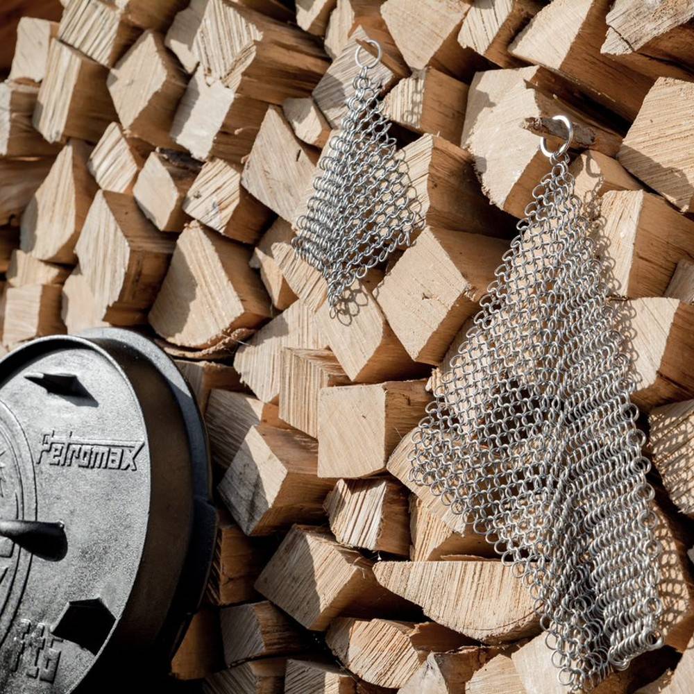 Pair of stainless steel chainmail scrubbers hanging in front of stacked firewood and a cast iron cookware lid, designed for cast and wrought iron cleaning, featured in Home & Decor, Household Cleaning, Cleaning Utensils and Gadgets, Best Sellers, and Promo Eligible collections.