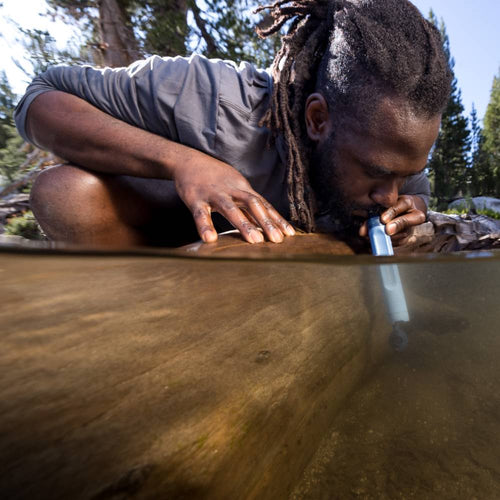 Man with dreadlocks using a portable water filter straw to drink directly from a natural freshwater stream in an outdoor camping setting surrounded by forest trees.