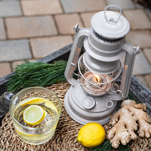 Handcrafted white galvanized oil lantern with durable thermal heat-resistant globe glowing softly on rustic woven mat alongside fresh lemon, ginger, and pine sprigs, ideal for Lamps & Lighting, Camping, and Emergency Supplies collections.
