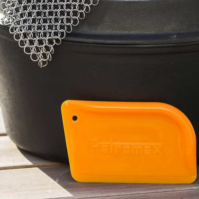 Orange Petromax pan scraper with a curved edge designed for gentle cleaning of cast iron cookware, shown next to a black Dutch oven, featured in Kitchen & Food and Cookware collections.