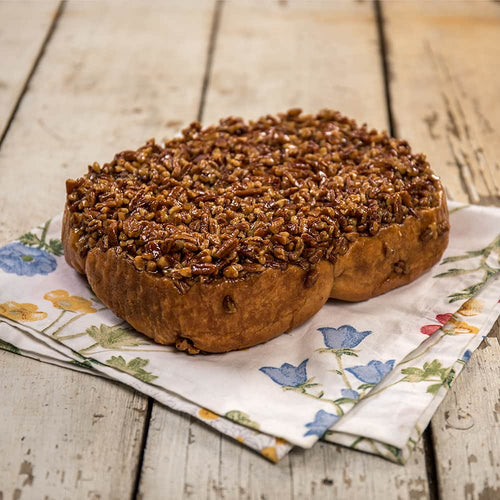 Large freshly baked pecan roll topped with chopped nuts and caramel glaze, resting on a floral cloth on rustic wooden surface, part of Kitchen & Food and USA Made collections