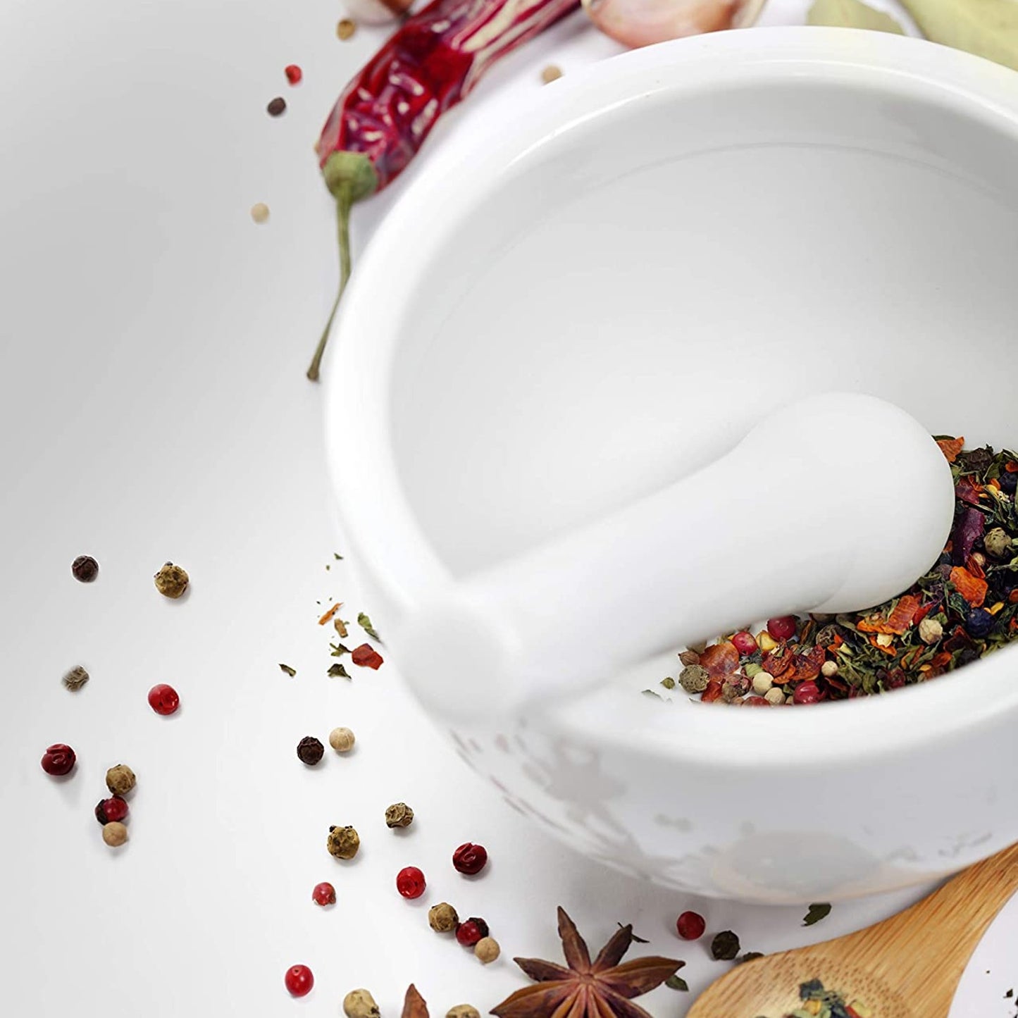 Close-up of a white porcelain mortar with pestle crushing mixed dried herbs and spices on a white surface, styled for Kitchen & Food and Cooking Helpers collections.