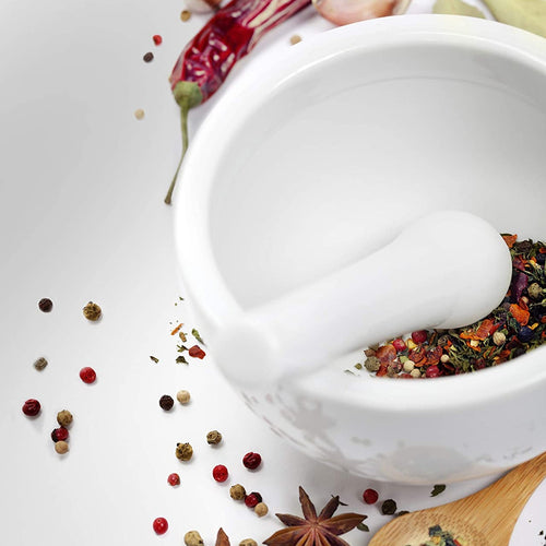 Close-up of a white porcelain mortar with pestle crushing mixed dried herbs and spices on a white surface, styled for Kitchen & Food and Cooking Helpers collections.