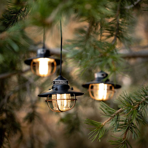 Three vintage-style Edison bulbs with protective black metal cages hanging from tree branches, emitting a warm glow, ideal for outdoor garden or camping lighting.