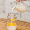 Clear plastic butter maker jar with yellow measuring markings being filled with heavy whipping cream from a glass pitcher, designed for easy homemade butter preparation in Farm, Garden & Tool and Kitchen & Food collections.