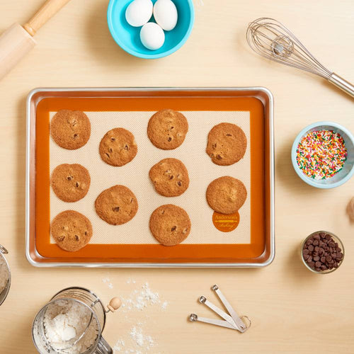 Metal baking sheet lined with a large non-stick silicone mat holding freshly baked chocolate chip cookies, surrounded by baking tools and ingredients for Kitchen & Food, Cookware, Baking Supplies collections