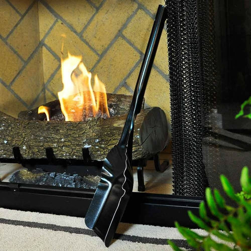 Black metal ash shovel with ergonomic handle leaning against a fireplace screen in front of a burning wood stove, handcrafted in USA, featured in Hearth Accessories and Stoves collections.