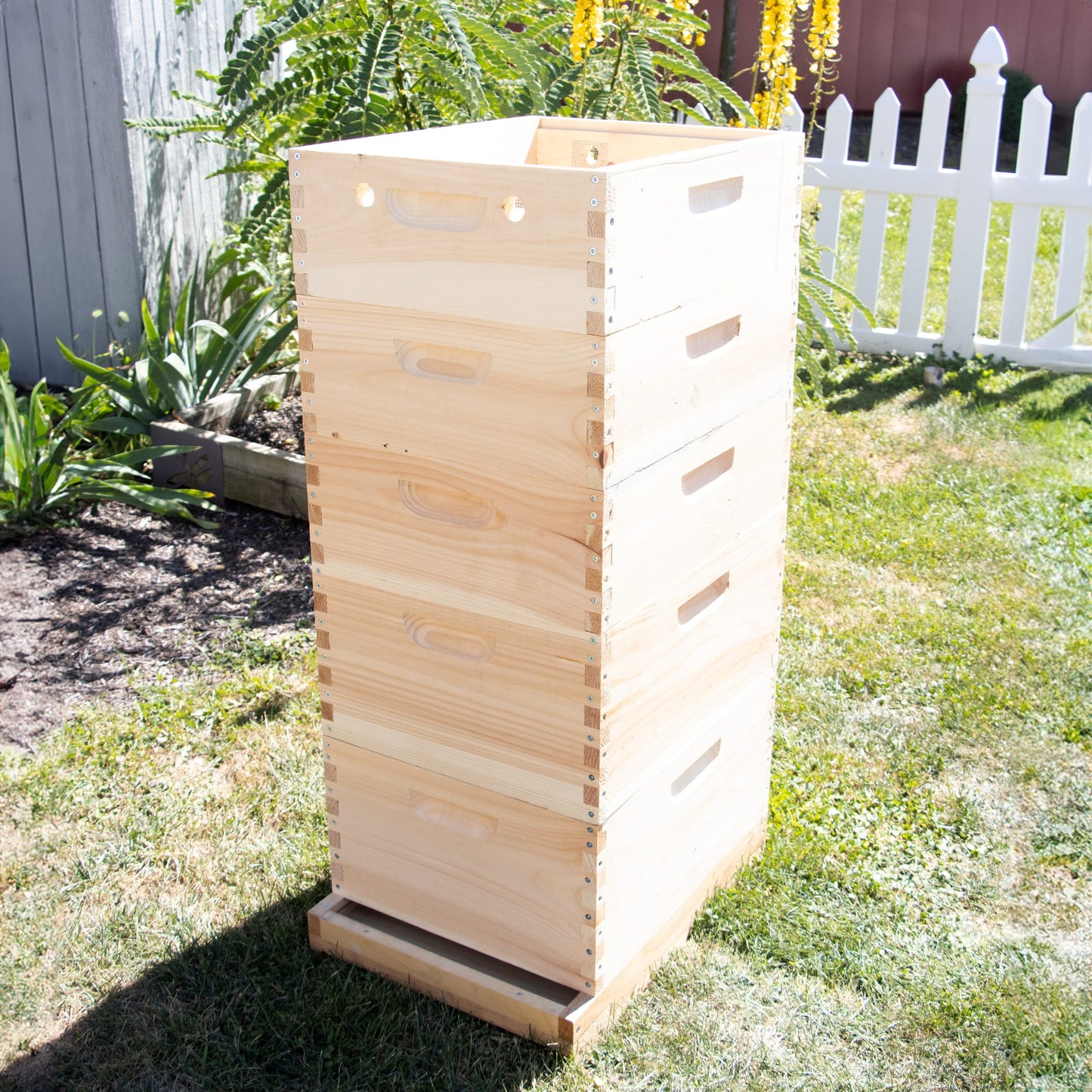 Stack of handmade wooden 10-frame quilt boxes for beekeeping, featuring dovetail joints and natural wood finish, set outdoors on grass near garden and white picket fence, Amish made and USA crafted.
