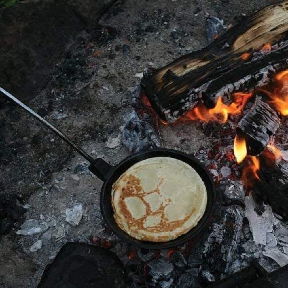 Cast iron skillet with long handle cooking a pancake over glowing campfire embers, showcasing durable cookware ideal for outdoor camping in Farm, Garden & Tool and Best Sellers collections.