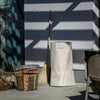 Beige ripstop fabric storage cover with full-length zipper and external fasteners, protecting a fire barrel, shown outdoors with wood basket and broom in Farm, Garden & Tool and Camping collections.