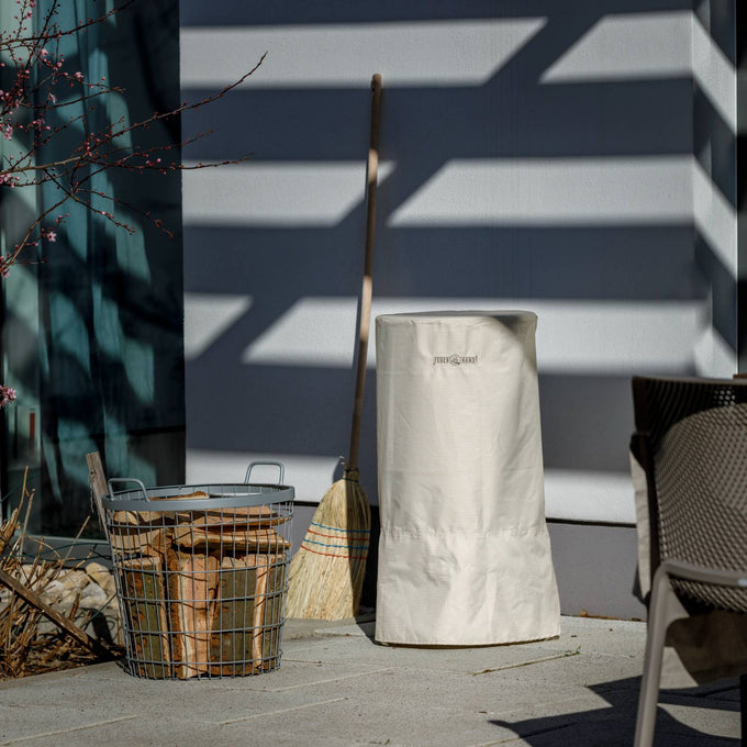 Beige ripstop fabric storage cover with full-length zipper and external fasteners, protecting a fire barrel, shown outdoors with wood basket and broom in Farm, Garden & Tool and Camping collections.