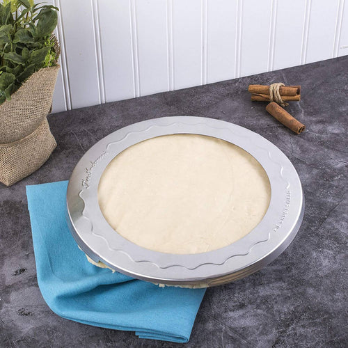 Aluminum pie crust shield protecting raw pie dough’s edges on a dark countertop with a blue cloth, cinnamon sticks, and a potted plant nearby, ideal for baking helpers and kitchen gadgets.