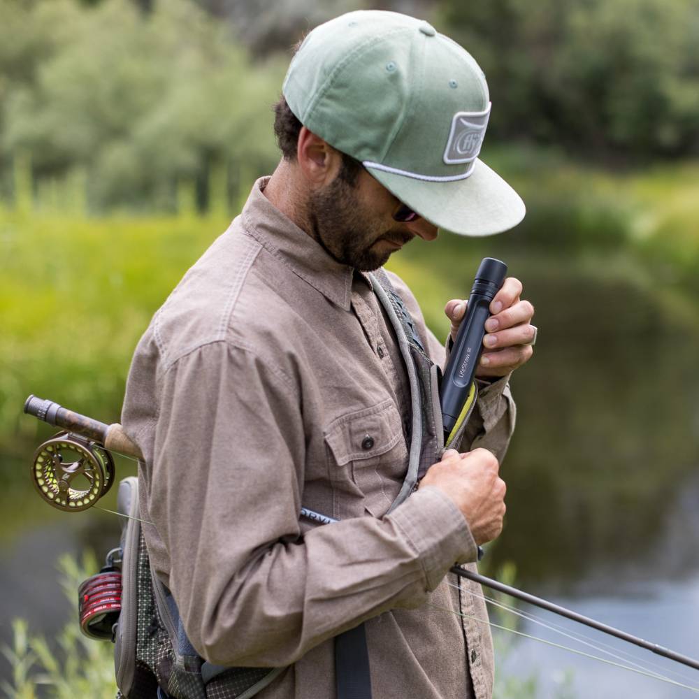 Man outdoors near a river in fishing gear placing a black LifeStraw Peak water filter straw into a pocket, preparing for camping or emergency water filtration use.