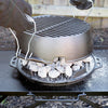 Gloved hands lifting a portable 12-inch cast iron grill lid with handles, showing hot charcoal briquettes underneath on a sturdy outdoor camping stove, handcrafted and made in USA.