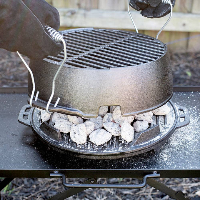 Gloved hands lifting a portable 12-inch cast iron grill lid with handles, showing hot charcoal briquettes underneath on a sturdy outdoor camping stove, handcrafted and made in USA.