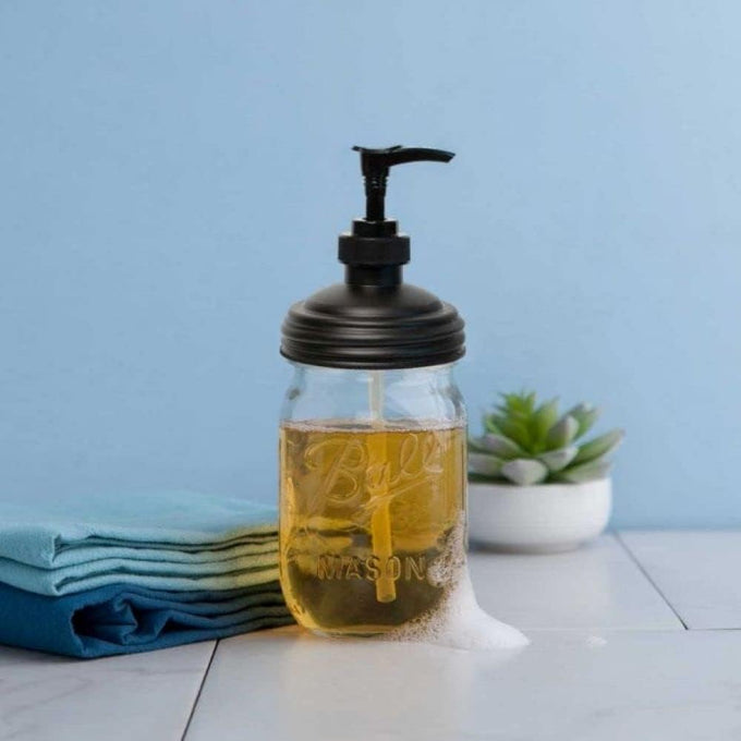 Clear glass regular-mouth mason jar filled with golden soap and topped with a durable black pump lid, set on a white surface with folded blue towels and a small green succulent, ideal for Health & Wellness, Home & Decor, Soaps and Soapmaking, and USA Made collections.
