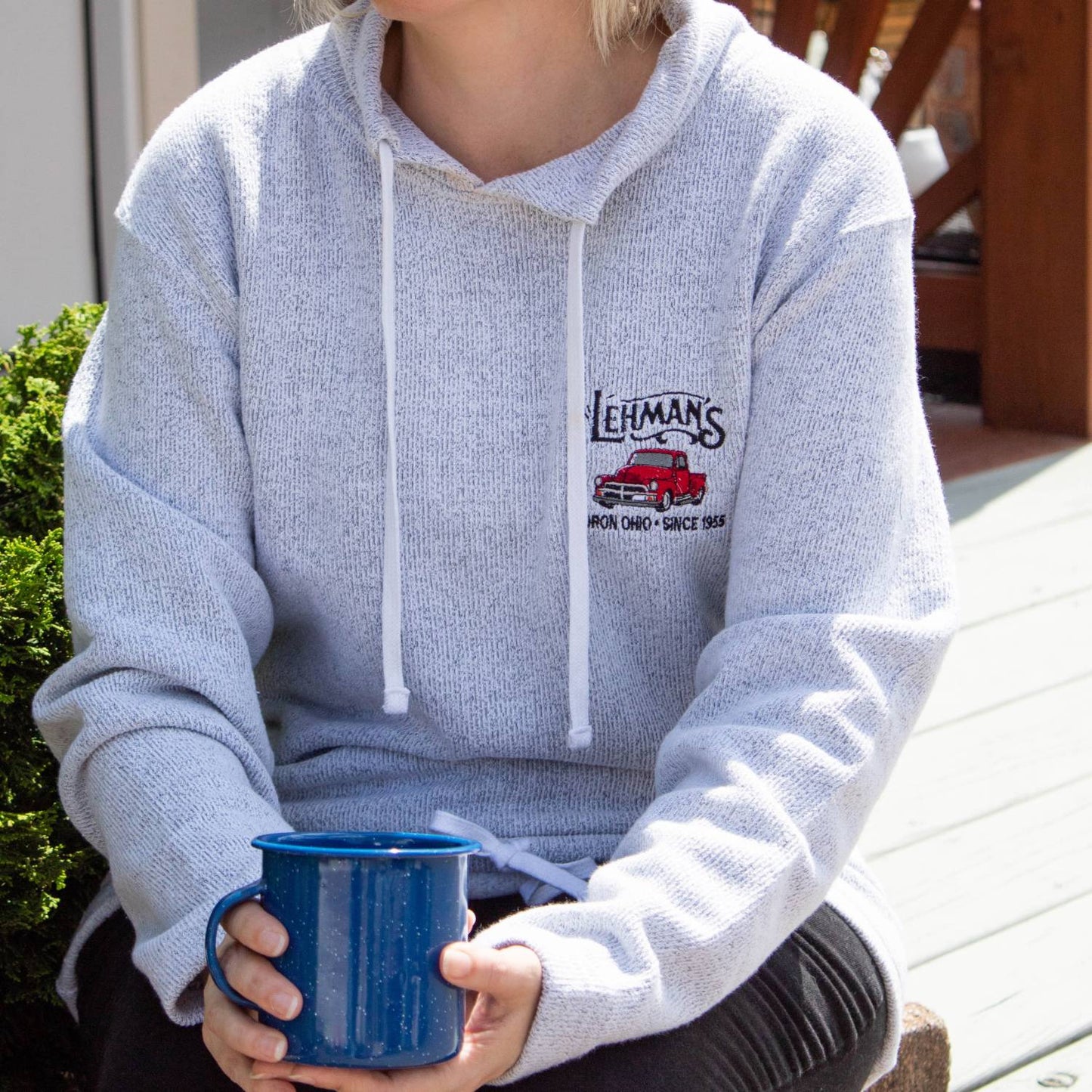 Person wearing a light gray handcrafted tunnel neck sweatshirt with embroidered classic red truck and Lehman’s logo, sitting outdoors holding a blue mug, featured in Health & Wellness, Clothing and Accessories, Best Sellers, and Promo Eligible collections.