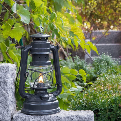 Handcrafted dark galvanized oil lantern with thermal heat-resistant SUPRAX globe, glowing softly while resting on garden stone near lush green foliage, ideal for camping, emergency supplies, and outdoor lighting in lamps & lighting collections.