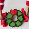 Cast iron cakelet pan with holiday-themed shapes filled with green and red batter, held by hands using red cloths, perfect for baking and seasonal kitchen use.