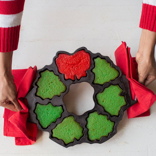 Cast iron cakelet pan with holiday-themed shapes filled with green and red batter, held by hands using red cloths, perfect for baking and seasonal kitchen use.
