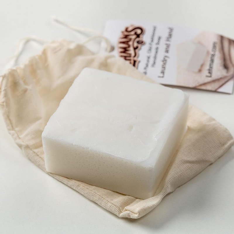 Handcrafted white natural laundry and hand soap bar made from organic coconut oil, resting on a cloth pouch with product information card, highlighting USA made, health, and grooming collections.