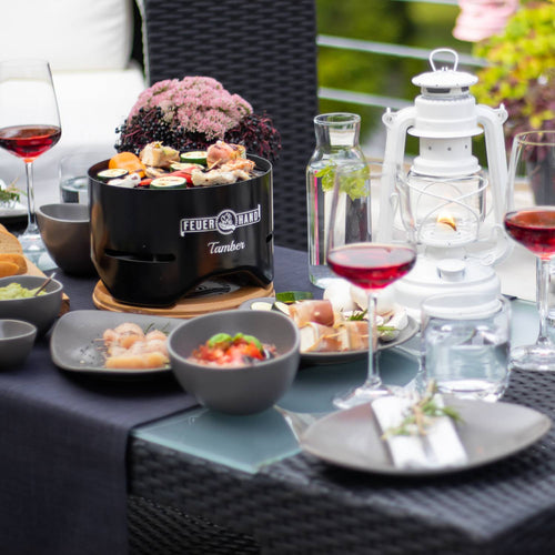 Black Feuerhand Tamber tabletop charcoal grill cooking assorted skewers, displayed on a glass patio table set with wine glasses, a white lantern, and outdoor dining accessories in Farm, Garden & Tool and Grilling collections.
