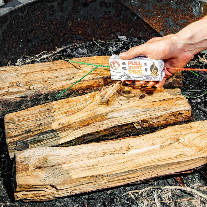 Hand holding a pull string fire starter pack with green ignition cords, placed over dry wood logs in a fire pit, ideal for Farm, Garden & Tool and Camping collections.