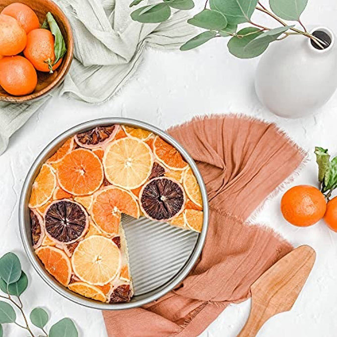 Round aluminized steel cake pan with fluted surface featuring a citrus-topped cake, styled with natural kitchen textiles and greenery, perfect for Kitchen & Food and Cookware collections.
