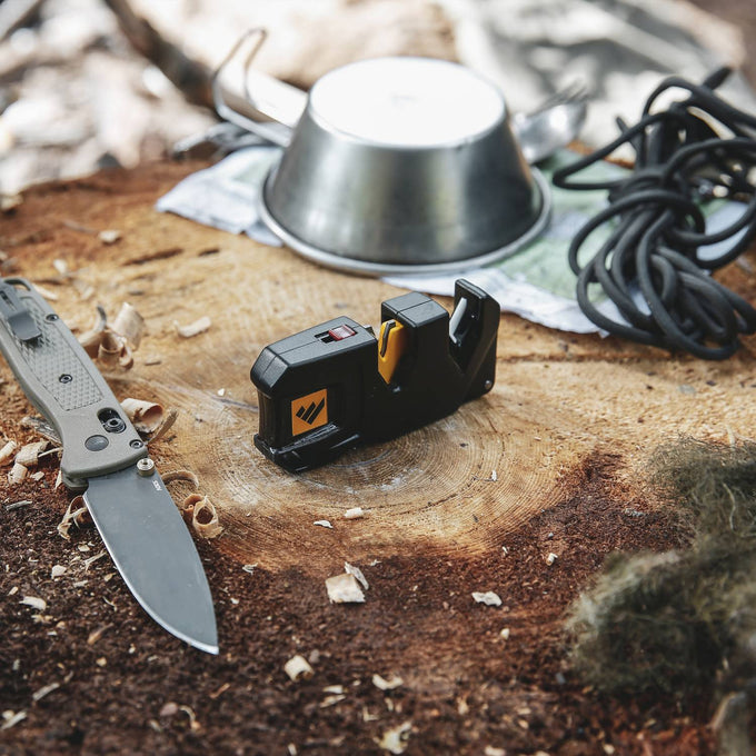 Compact black and yellow handheld knife sharpener with ergonomic design, shown on a rustic wood surface alongside a folding knife and outdoor gear, ideal for farm, garden, kitchen, and sharpening tool collections.