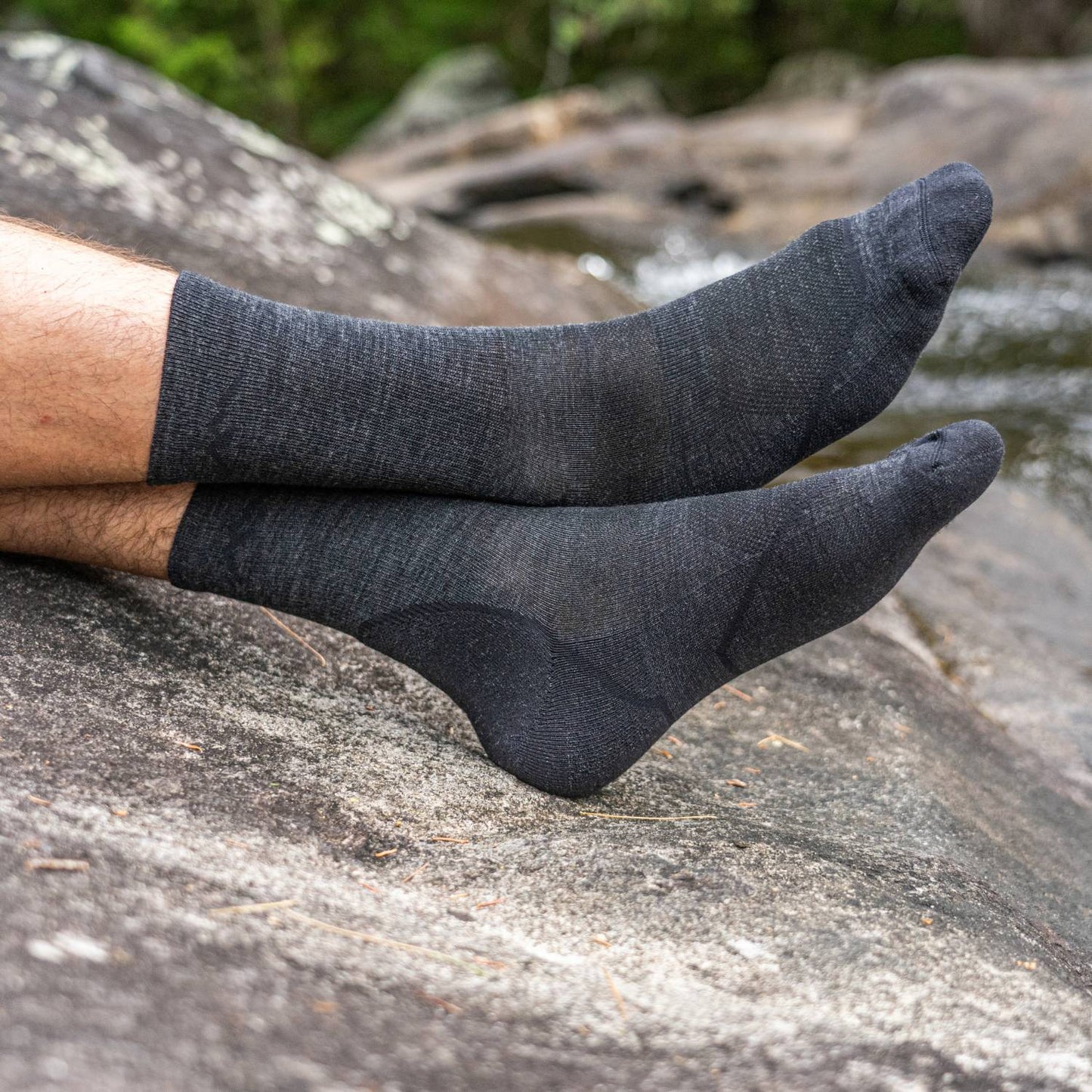 Pair of dark gray lightweight micro crew socks with smooth ribbing on a person's feet resting on a natural rock surface, ideal for camping, health & wellness, and outdoor activities.