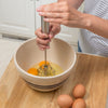 Hand using a stainless steel whisk to blend eggs in a ceramic mixing bowl on a wooden countertop, suitable for Kitchen & Food, Kitchen Gadgets, Cooking and Baking Helpers, Best Sellers, and Promo Eligible collections.