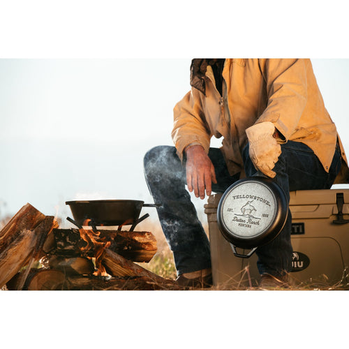 Person wearing outdoor gloves and jacket holding a handcrafted cast iron Bucking Bronco combo cooker near a campfire with wood logs and cooking pot, featured in Kitchen & Food, Cast Iron, USA Made, and Best Sellers collections.