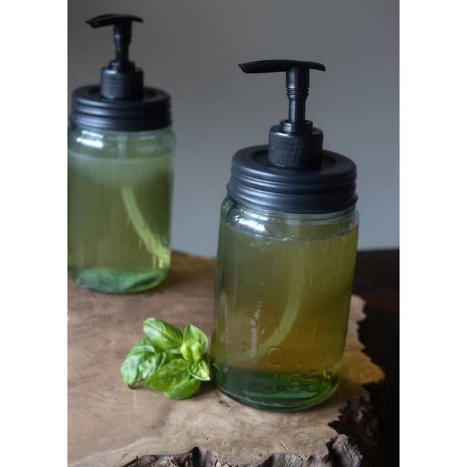 Black pump lids with dispensing tubes fitted on clear regular-mouth glass jars filled with liquid soap, displayed on a rustic wooden surface with fresh basil, highlighting USA made and eco-friendly Health & Wellness, Home & Decor, and Household Helpers collections.
