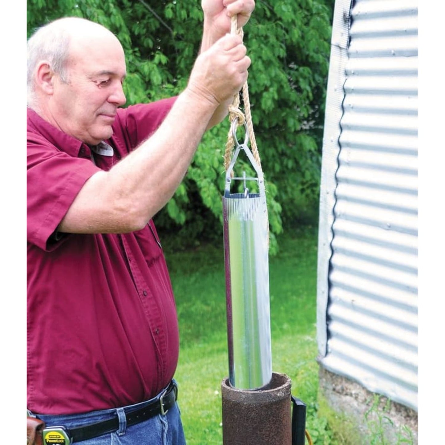 Man lifting a handcrafted galvanized steel well bucket by rope from a metal well casing outdoors, suitable for farm, garden, emergency, and best sellers collections.