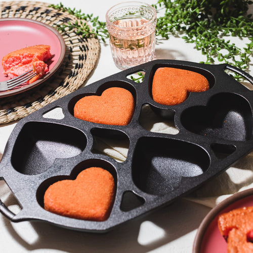 Cast iron mini cake pan featuring six heart-shaped molds with a naturally seasoned surface, holding freshly baked orange-red heart cakes, perfect for Kitchen & Food, Cookware, Cast Iron, Baking Supplies, and USA Made collections.
