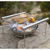Compact stainless steel grill with cooking grate elevated above glowing charcoal, grilling sausages, meat, and vegetable skewers outdoors, ideal for camping and backyard use.