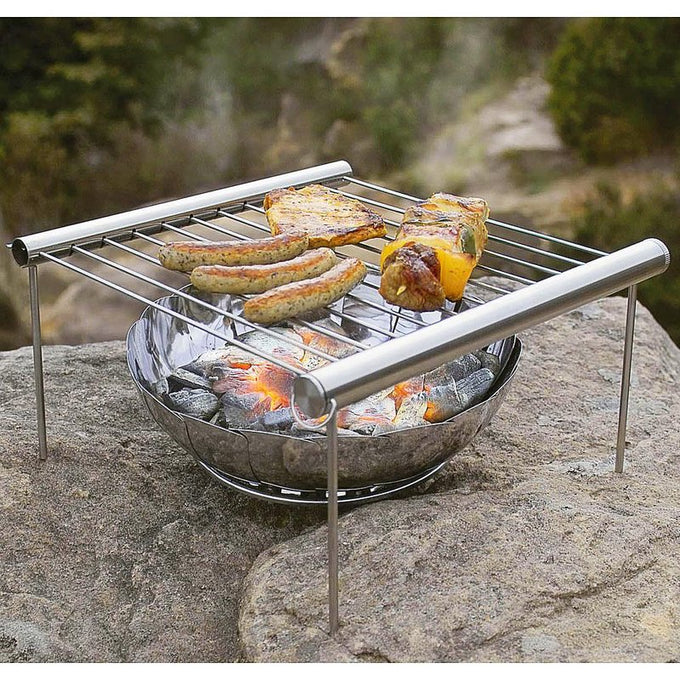 Compact stainless steel grill with cooking grate elevated above glowing charcoal, grilling sausages, meat, and vegetable skewers outdoors, ideal for camping and backyard use.