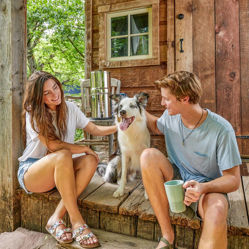 Young couple sitting on rustic wooden porch with a border collie, next to a compact stainless steel gravity water filter system, perfect for emergency supplies and travel needs.