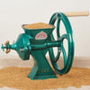 Cast iron grain mill with a green finish and wooden handle, grinding whole grains in a kitchen setting, featured in baking supplies and cookware collections.