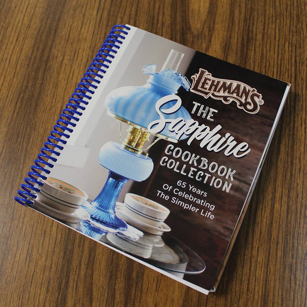 Hardcover cookbook with a blue spiral binding, featuring a vintage blue glass oil lamp and teacups on the cover, celebrating 65 years of simple living recipes in a unique, USA-made collection.