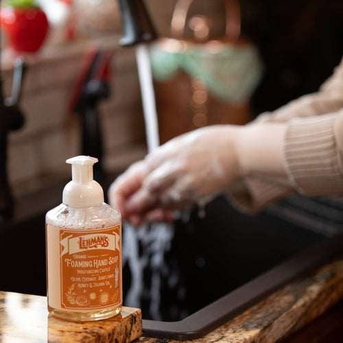 Clear pump bottle of handcrafted moisturizing foaming hand soap with natural oils and honey, made in USA, positioned on a kitchen counter near someone washing hands, ideal for Health & Wellness and Grooming collections.