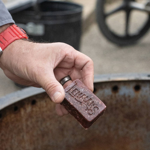 Handcrafted reddish-brown fire starter block embossed with Lehman’s logo, held above a metal fire ring, designed for easy, non-toxic fire lighting in stoves, camping, and hearth accessories from USA Made collection.