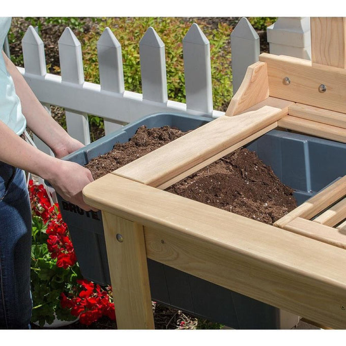 Close-up of a handcrafted Amish-made outdoor potting table made of smooth unfinished cypress wood, featuring a removable Rubbermaid tote filled with soil, set against a white picket fence and garden flowers.