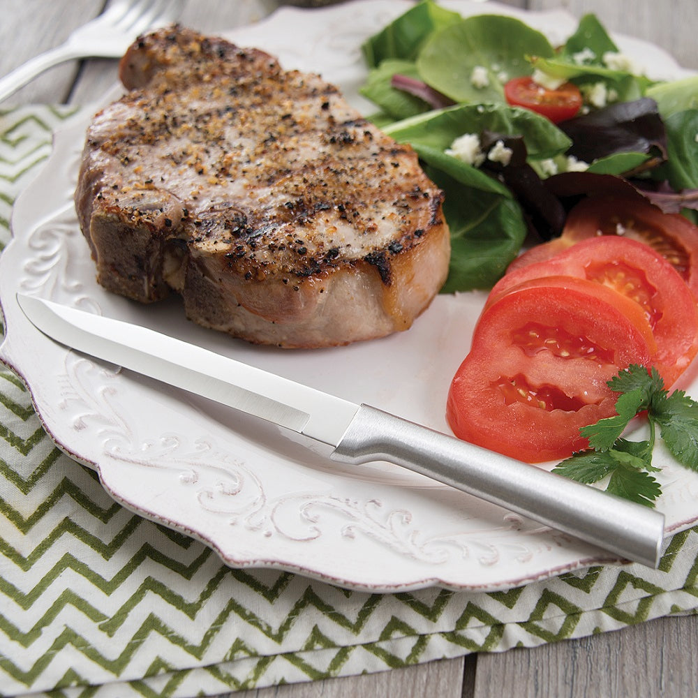 Rada steak and utility knife with solid satin-finished recycled aluminum handle, shown beside a seasoned grilled pork chop and fresh tomato salad, handcrafted in USA.