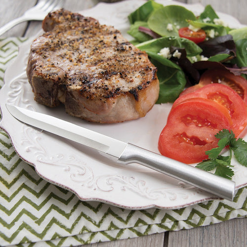 Rada steak and utility knife with solid satin-finished recycled aluminum handle, shown beside a seasoned grilled pork chop and fresh tomato salad, handcrafted in USA.