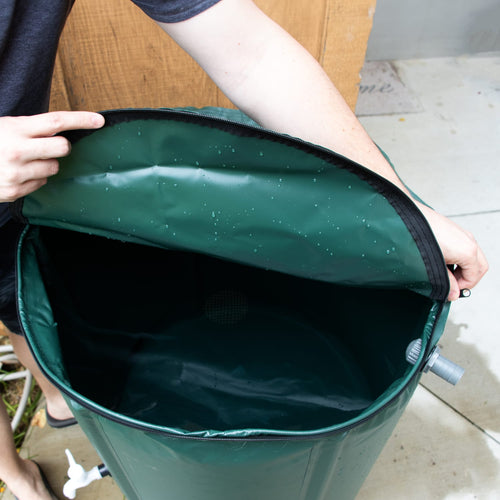 Person lifting the mesh-covered lid of a collapsible green rain barrel with a gray spigot on the side, featured in Farm, Garden & Tool, Garden Accessories, Lawncare, Best Sellers, and Promo Eligible collections.