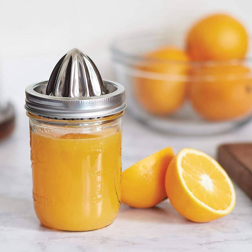 Sturdy stainless steel citrus juicer lid attached to a wide mouth jar filled with fresh orange juice, shown with halved oranges and a bowl of whole fruit on a kitchen countertop.